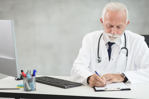 Older-male-doctor-with-white-hair-and-beard-writing-on-a-clipboard-at-his-desk.png